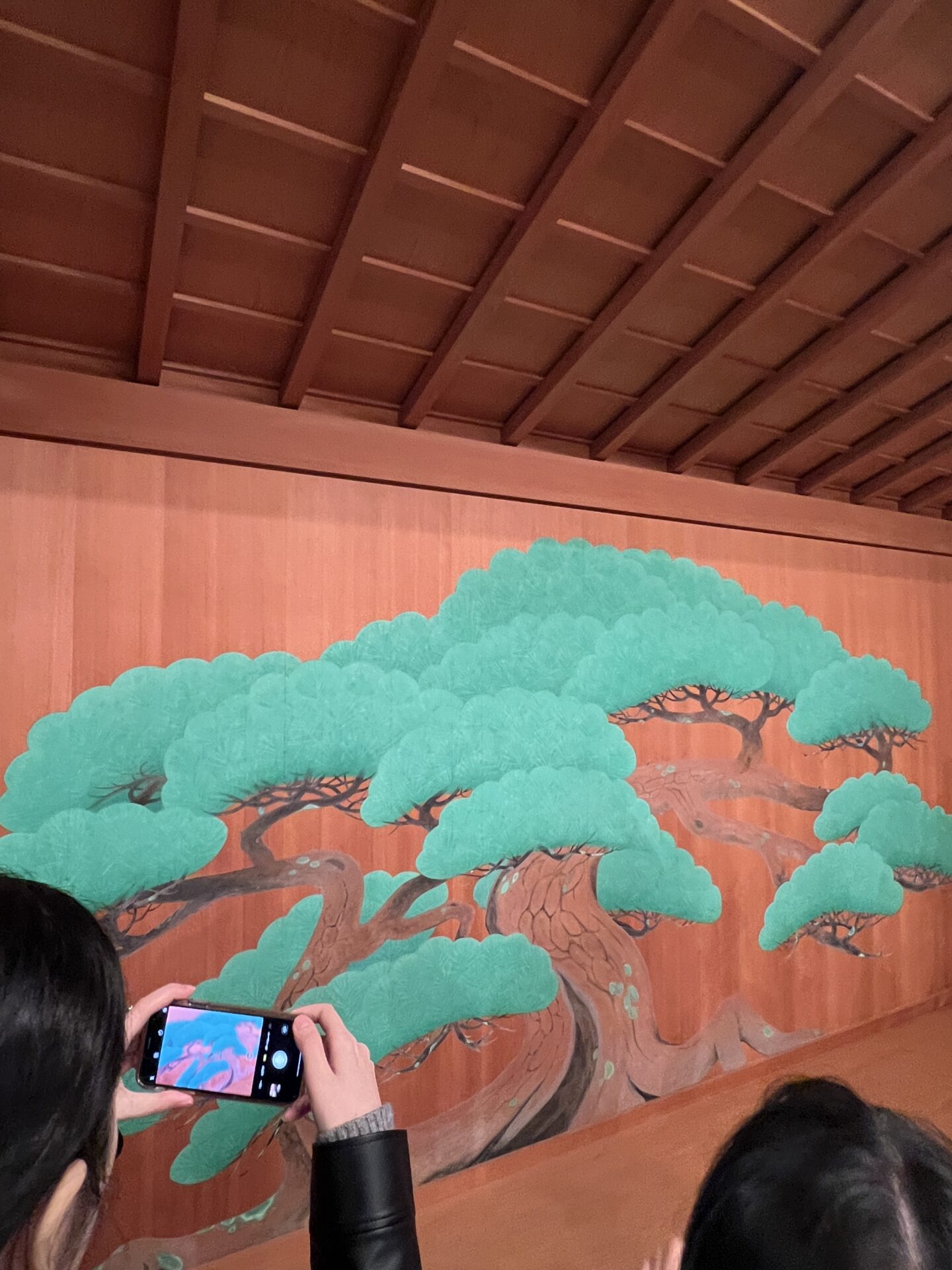 The iconic painted pine tree backdrop of a Japanese Noh theater stage, with someone in the audience snapping a photo on their phone.