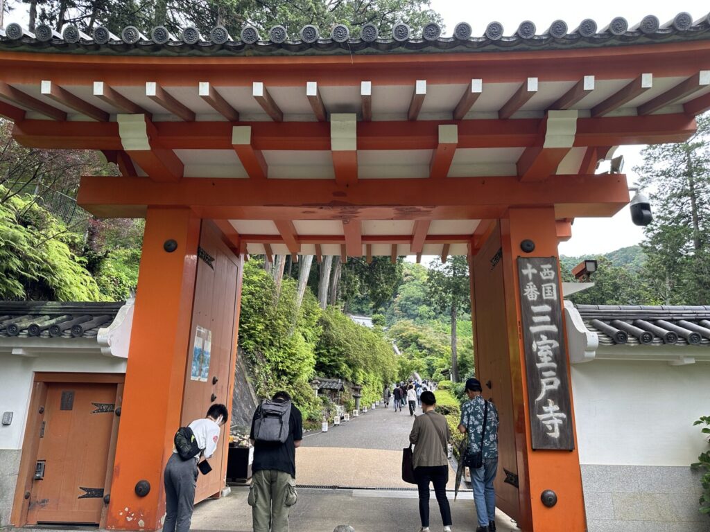 Walking through the striking vermilion main gate of Mimurotoji temple, with visitors filing in along a tree-lined path that climbs into the lush green hillside beyond.